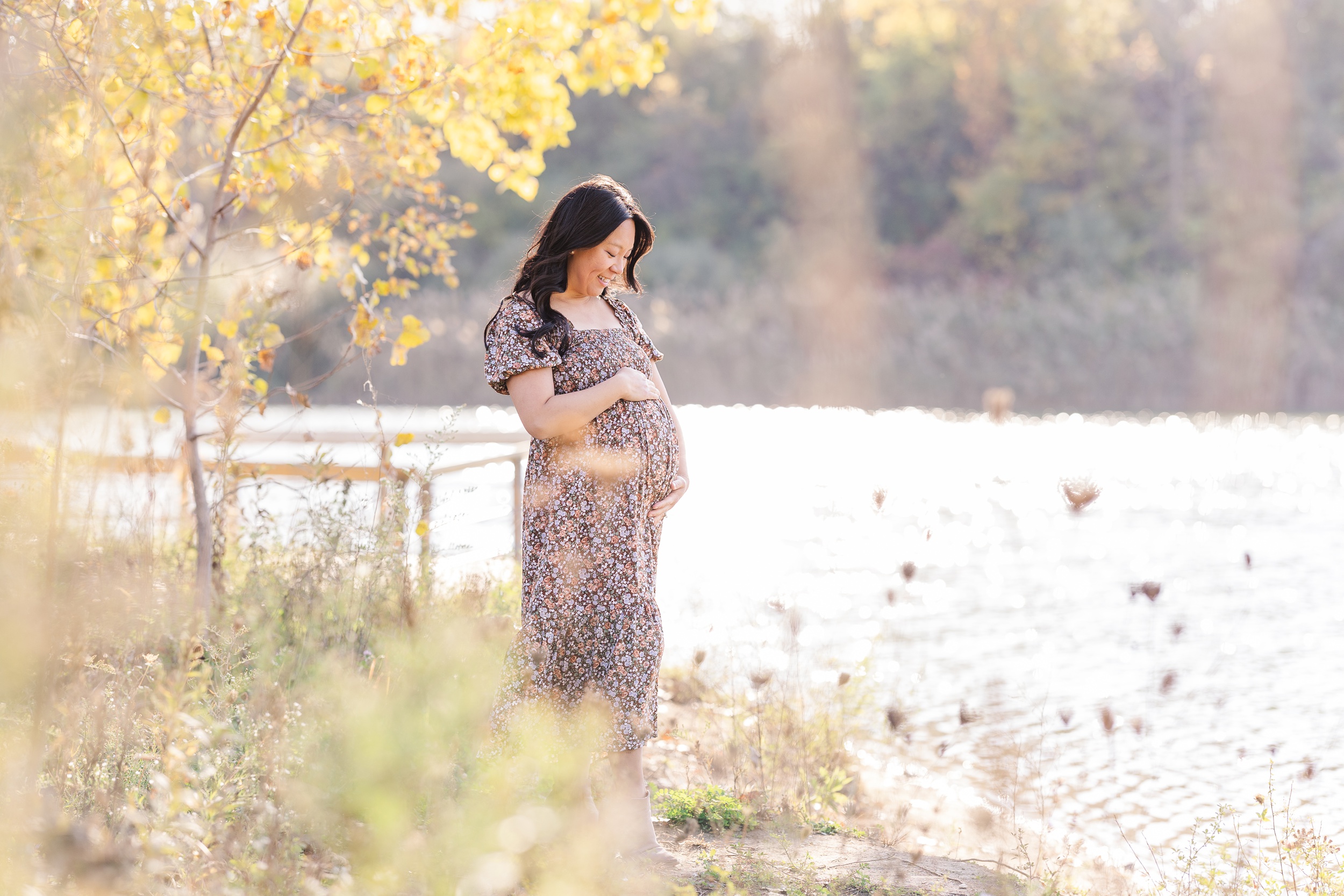 A smiling mother to be looks down to her bump in her hands while standing on a lakeshore at sunset