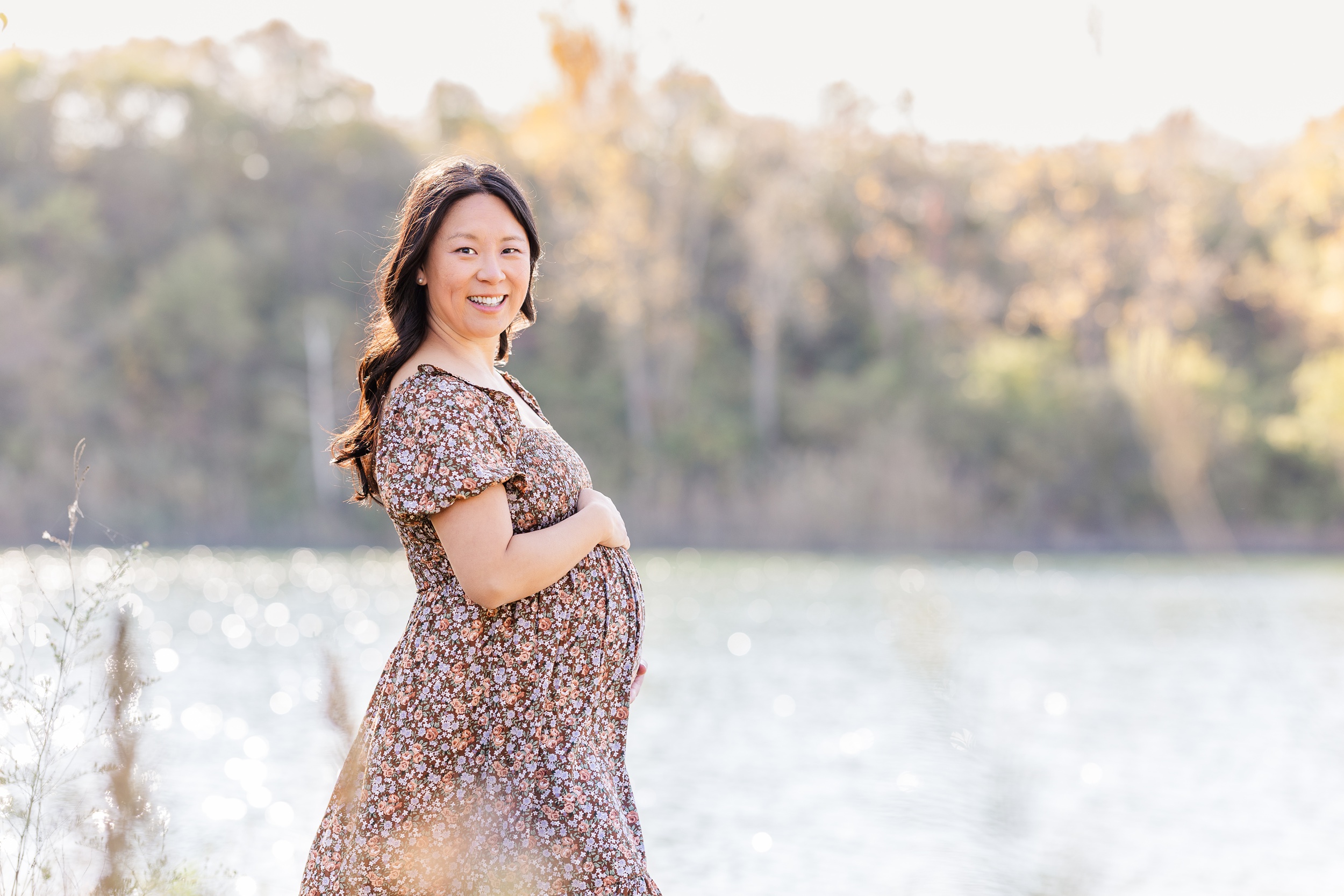 A mom to be in a floral print dress holds her bump while smiling and standing lakeside after meeting ann arbor doulas