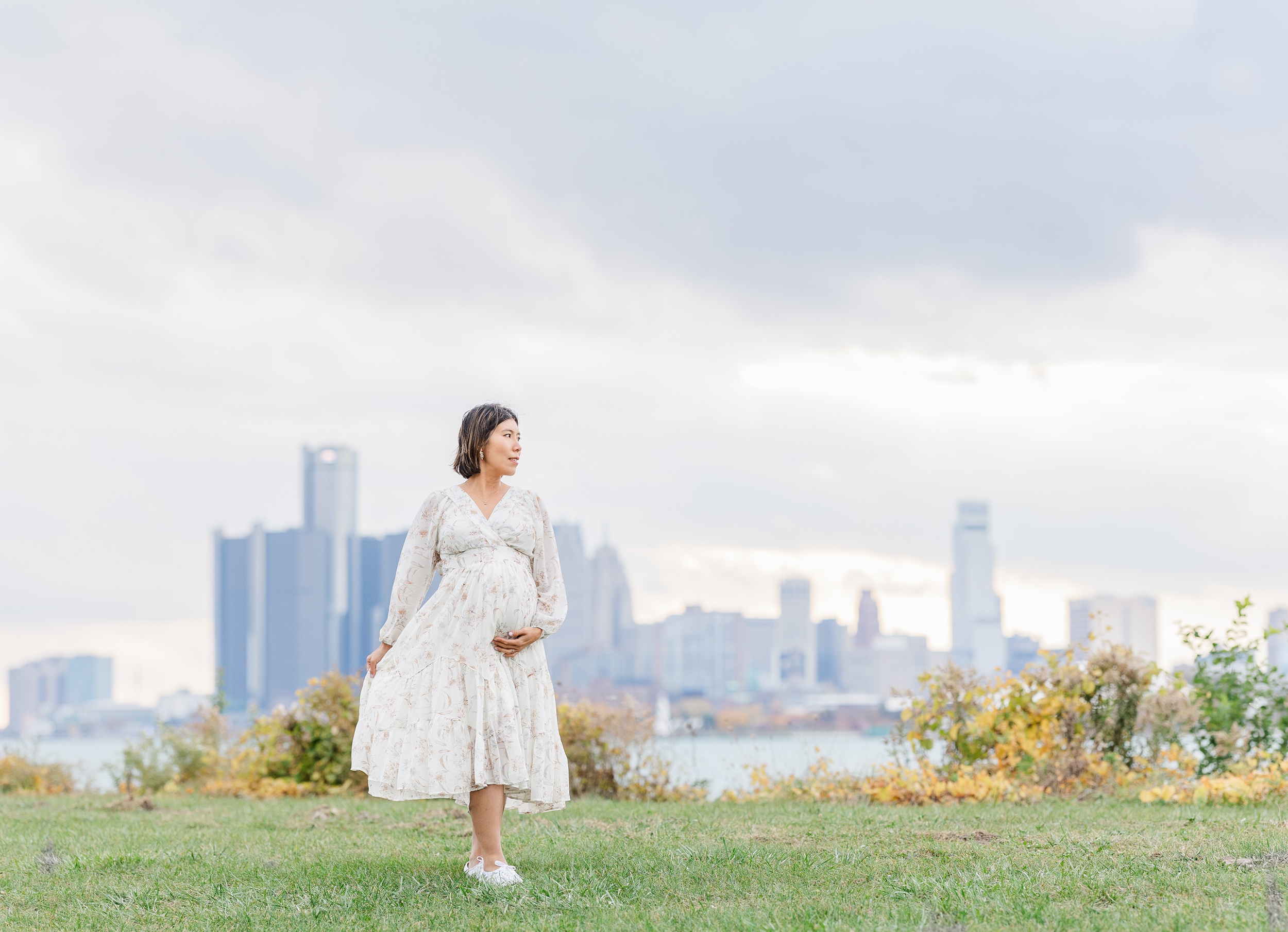 A happy mom to be explores a lakeside park at sunset in a white floral print dress after some ann arbor prenatal yoga