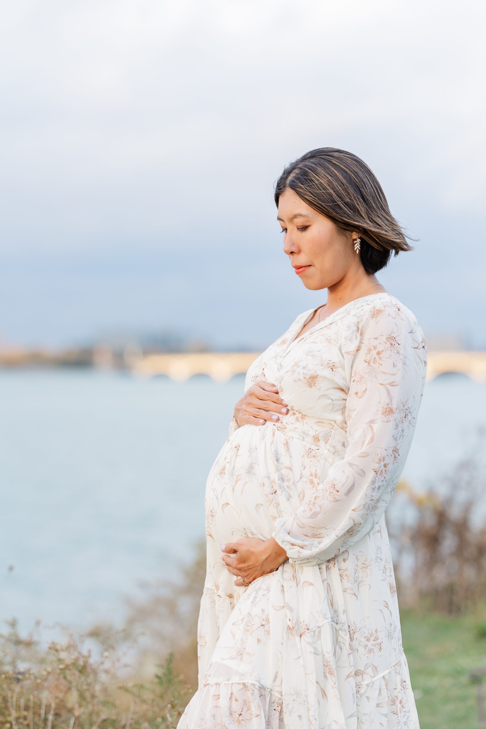 An expecting mother in a white floral dress stands by the water at sunset holding her bump and looking down to it after some ann arbor prenatal yoga