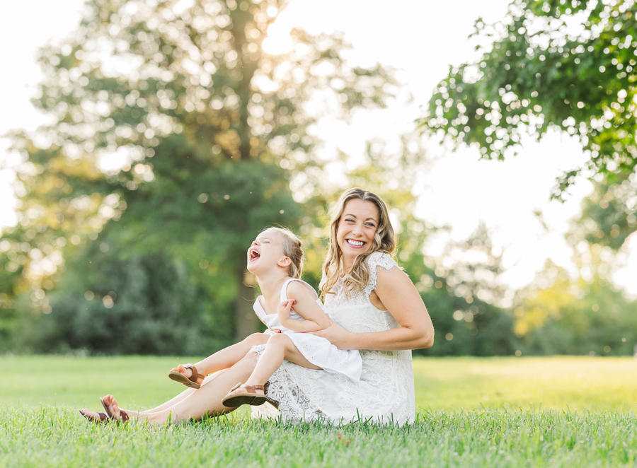 A laughing mom plays with her toddler daughter in a park lawn at sunset as they wear white dresses and sandals after visiting ann arbor toy stores