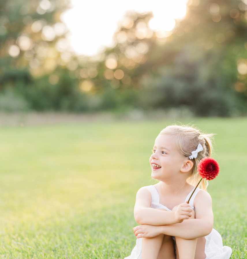 A smiling young girl in a white dress sits in a park lawn holding a large red flowers at sunset after visiting ann arbor toy stores