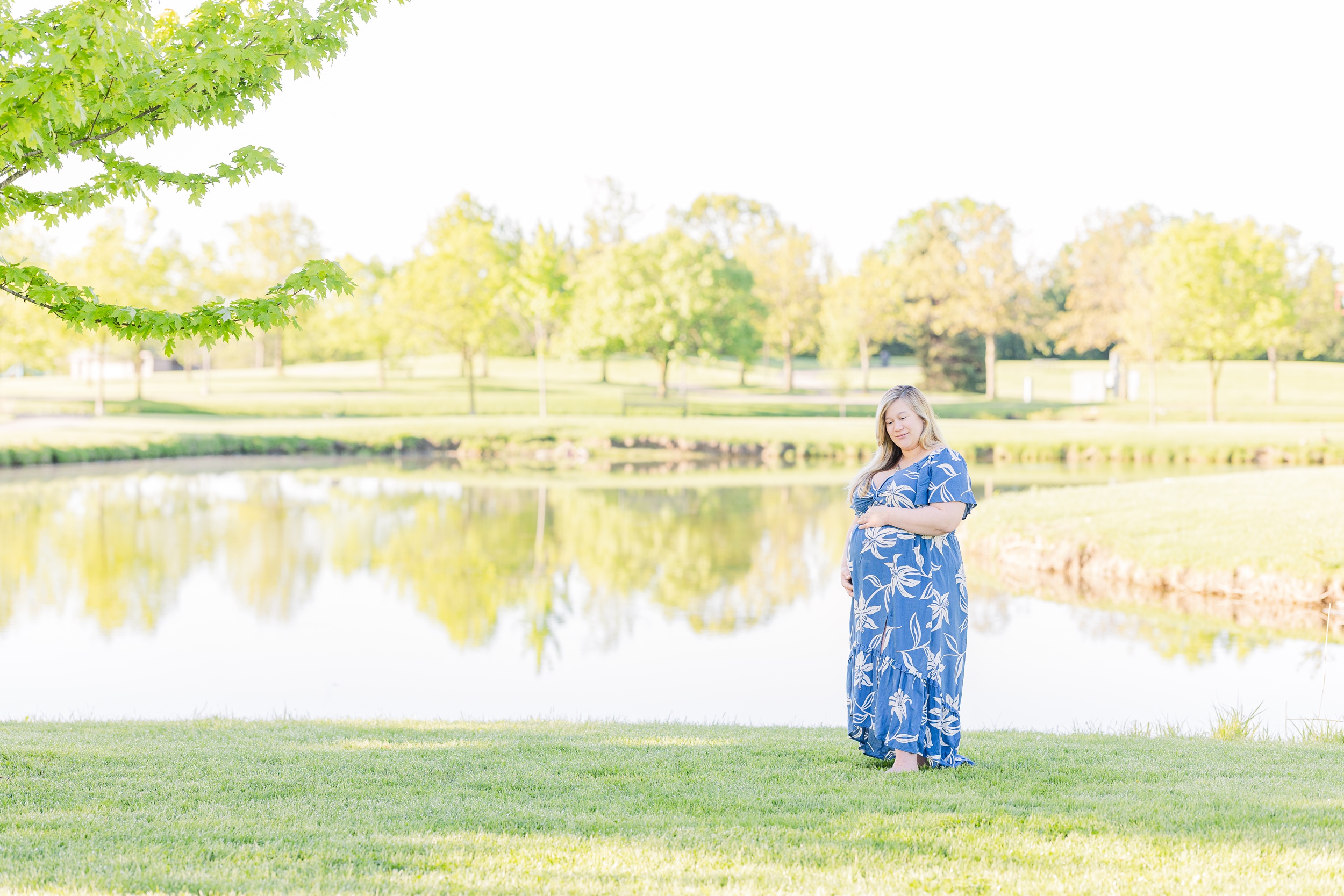 A smiling mother to be stands by a park pond in the lawn wearing a blue maternity dress after visiting birthing centers in Ann Arbor