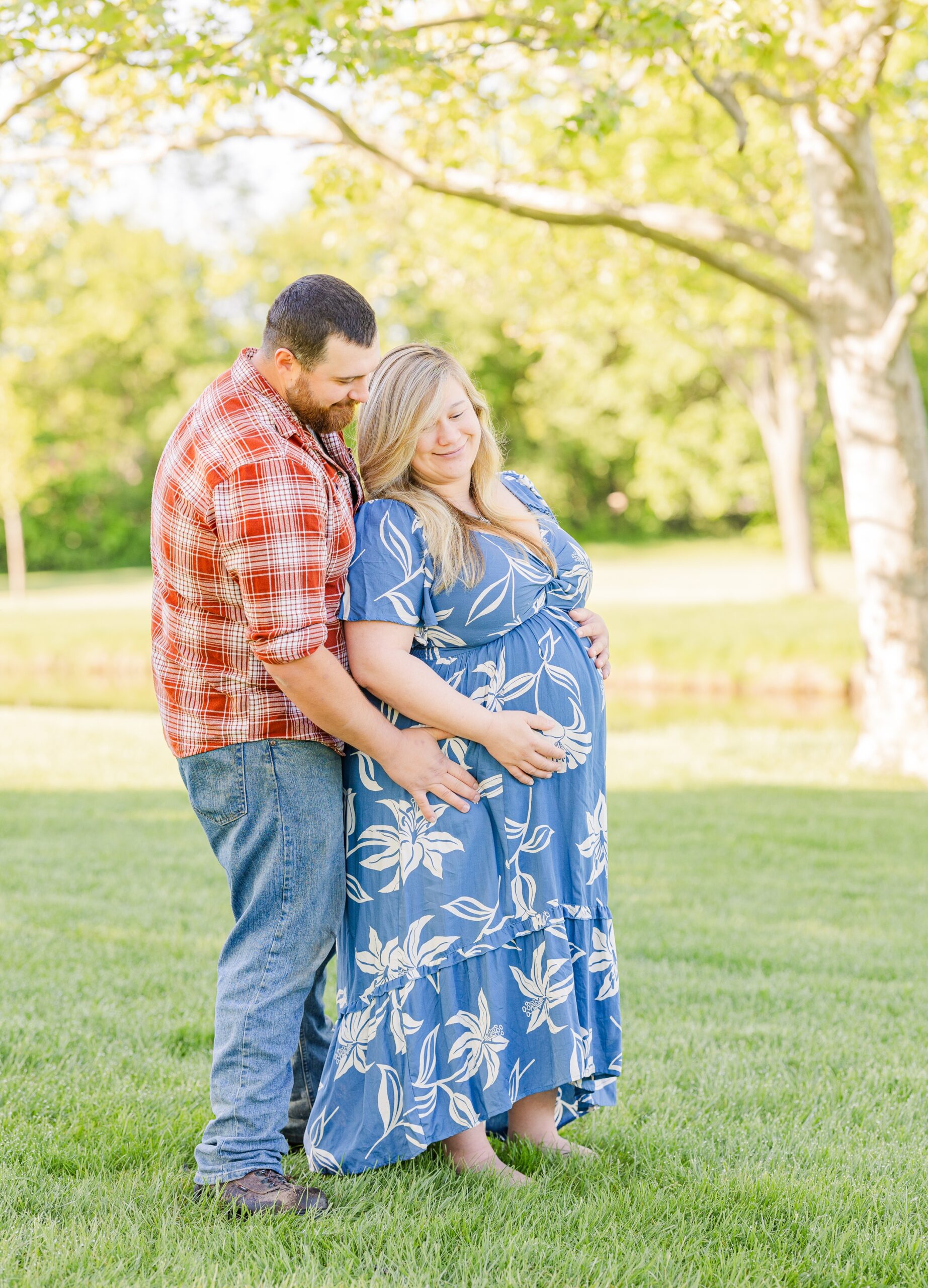 Happy expecting parents stand in a park lawn with hands on the bump after visiting birthing centers in Ann Arbor