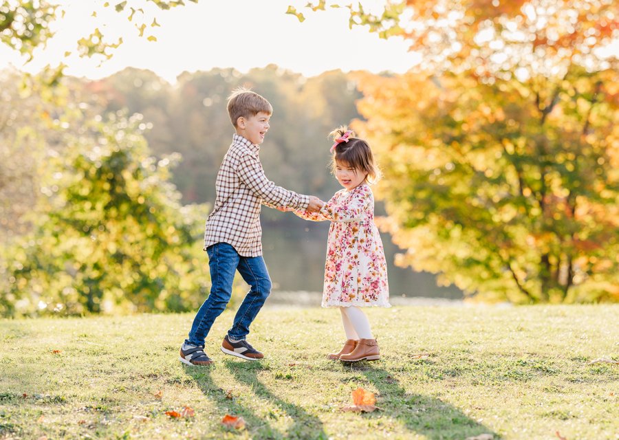 Happy toddler brother and sister dance and play in a field by a lake at sunset