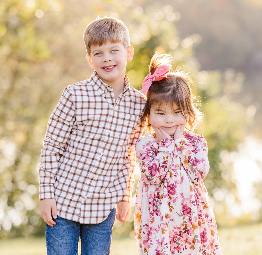 A happy young boy stands in jeans in a park at sunset smiling while his baby sister in a pink dress makes a silly face after visiting daycares in ann arbor