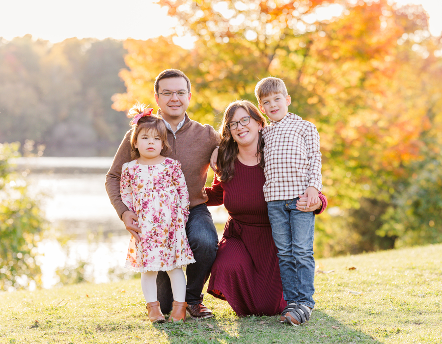 A happy mom and dad kneel by a lake in a park with their toddler son and daughter after visiting daycares in ann arbor