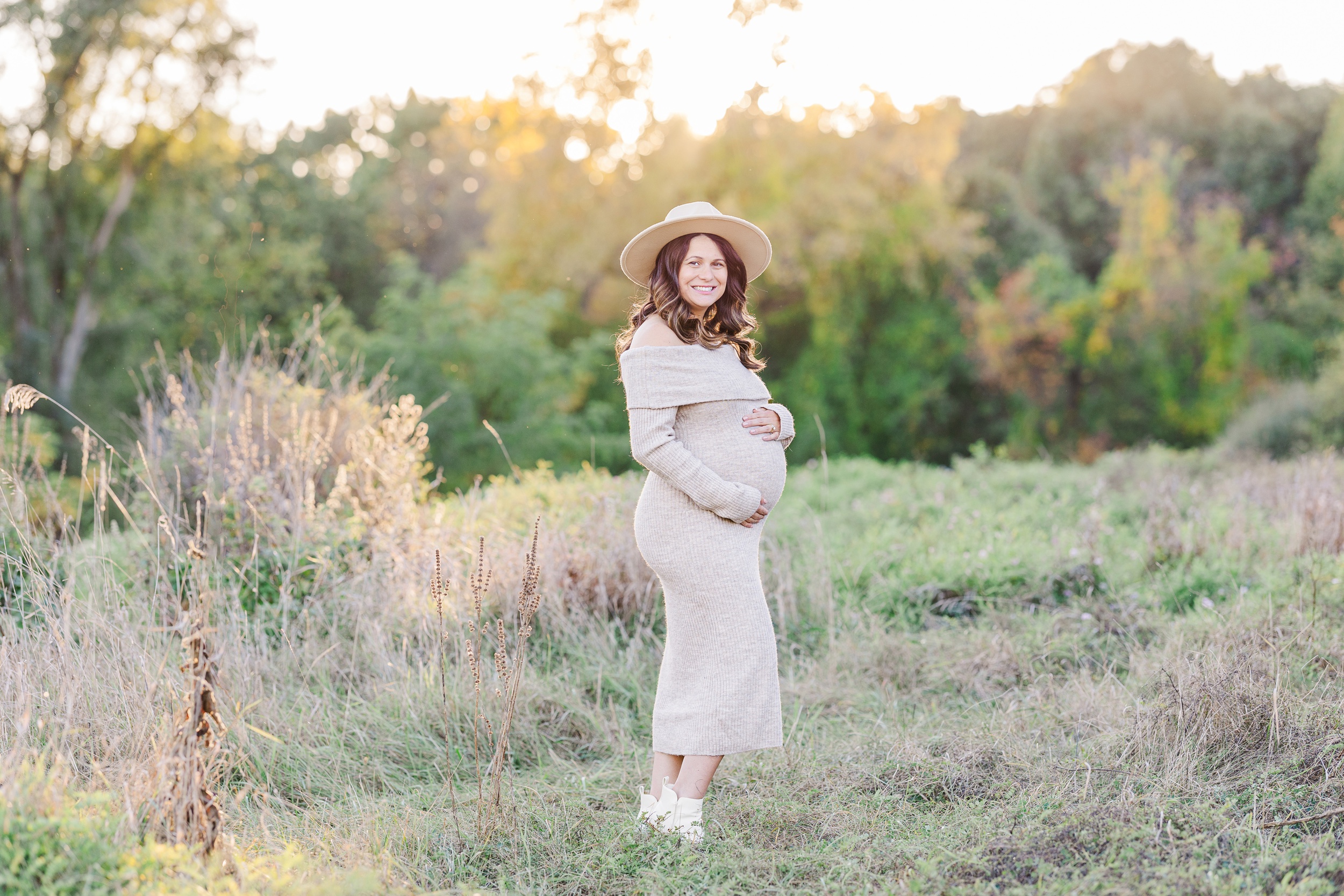 A smiling mom to be in a knit maternity gown stands in a field at sunset after meeting midwives in ann arbor