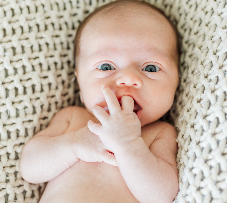 A newborn baby lays in a woven blanket with a finger in its mouth and eyes open