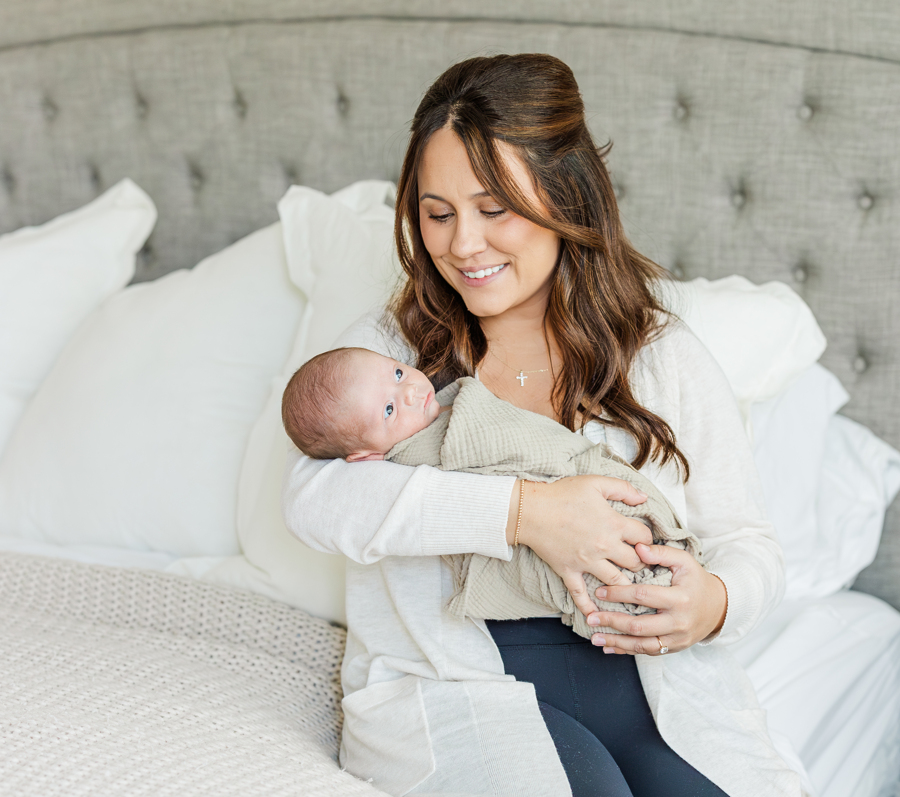 A smiling new mom in a white cardigan sits on the edge of a bed holding her awake newborn baby in her arms after meeting nannies in ann arbor