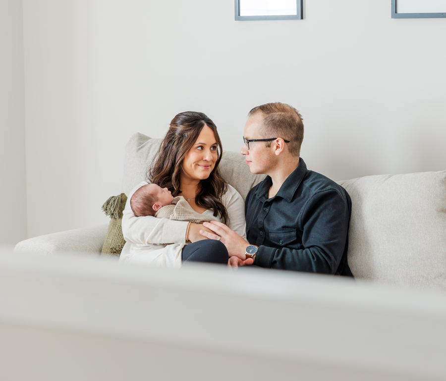 Happy new parents sit on a couch smiling at each other while mom holds their sleeping newborn after meeting nannies in ann arbor