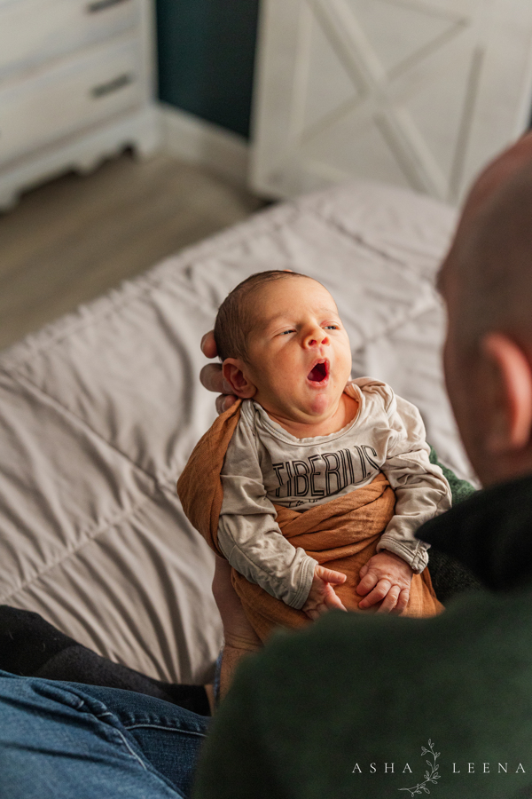 A newborn baby yawns while laying in dad's hands as he sits on a bed after meeting a nanny in Ann Arbor