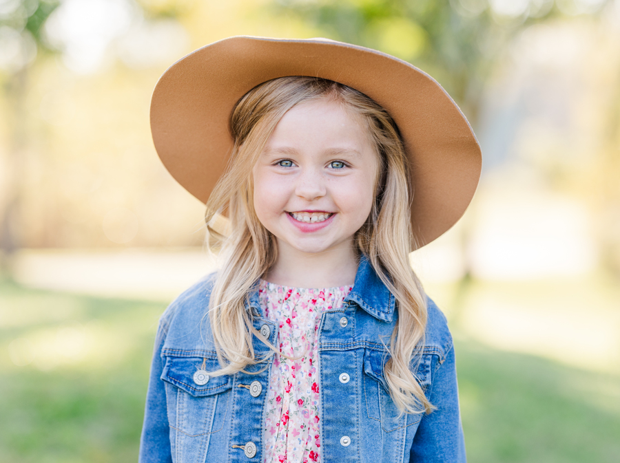 A happy young girl in a denim jacket and large hat smiles while standing in a park