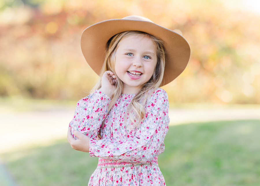 A smiling toddler girl in a pink dress and big hat laughs in a park after visiting a pediatric dentist in Ann Arbor