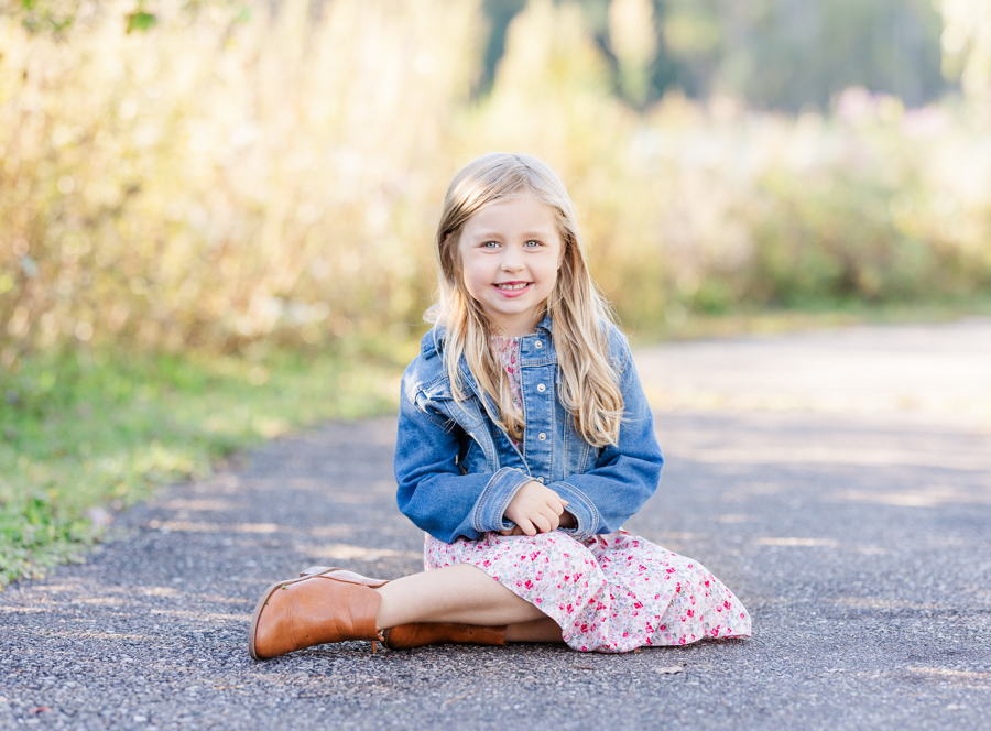 A toddler girl sits in the sidewalk of a park at sunset in a pink dress and denim jacket before visiting a pediatric dentist in Ann Arbor