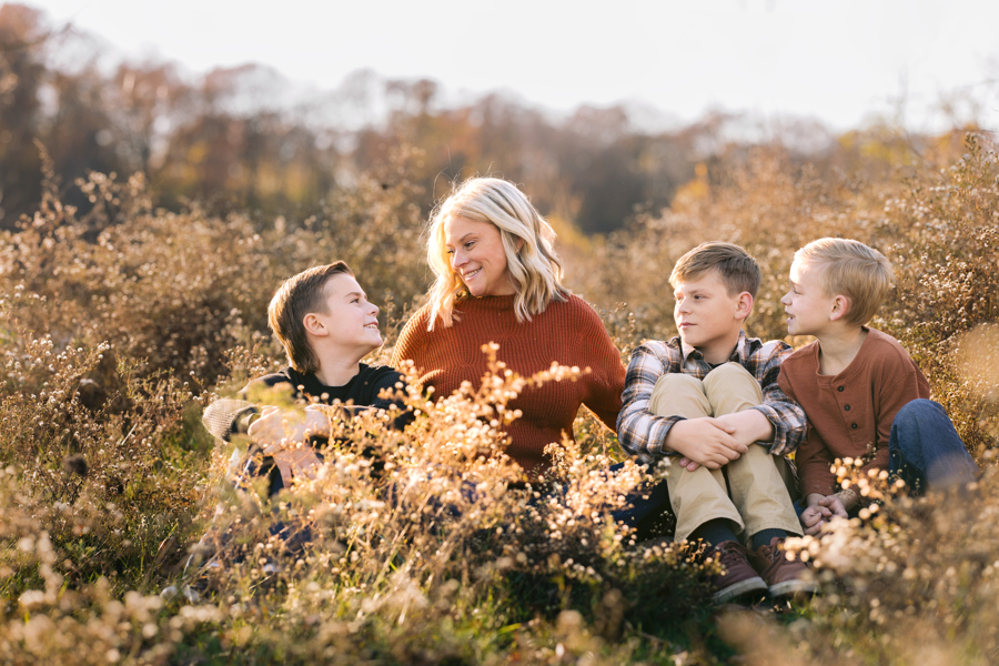 A happy mom smiles at one of her three young sons sitting in some tall golden grasses at sunset after visiting pediatricians in Ann Arbor