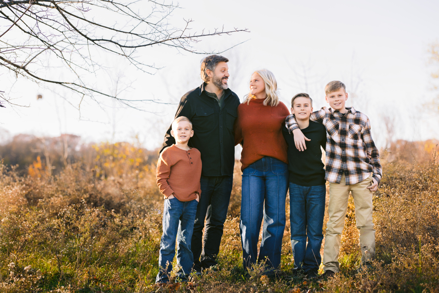 Happy mom and dad smile at each other while standing in a pasture with their three young sons after visiting pediatricians in ann arbor