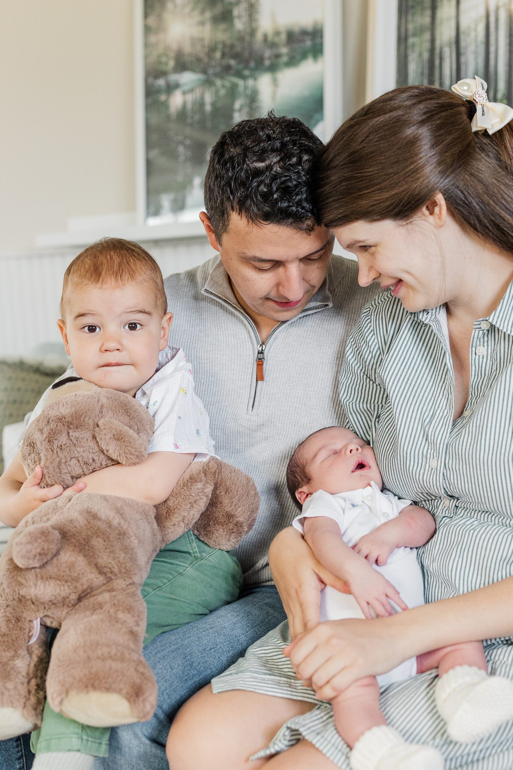 Happy mom and dad sit on a couch smiling with their toddler son and newborn baby in their laps after meeting a postpartum doula in Ann Arbor