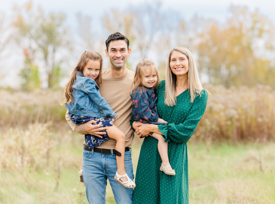 A smiling mother and father stand in a field of tall grass holding their toddler daughters on their hips