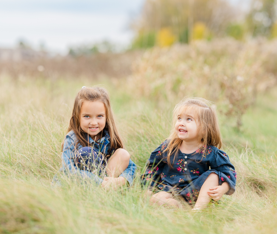 Toddler sister girls in blue dresses make silly faces while sitting in tall grass together during things to do in ann arbor with toddlers