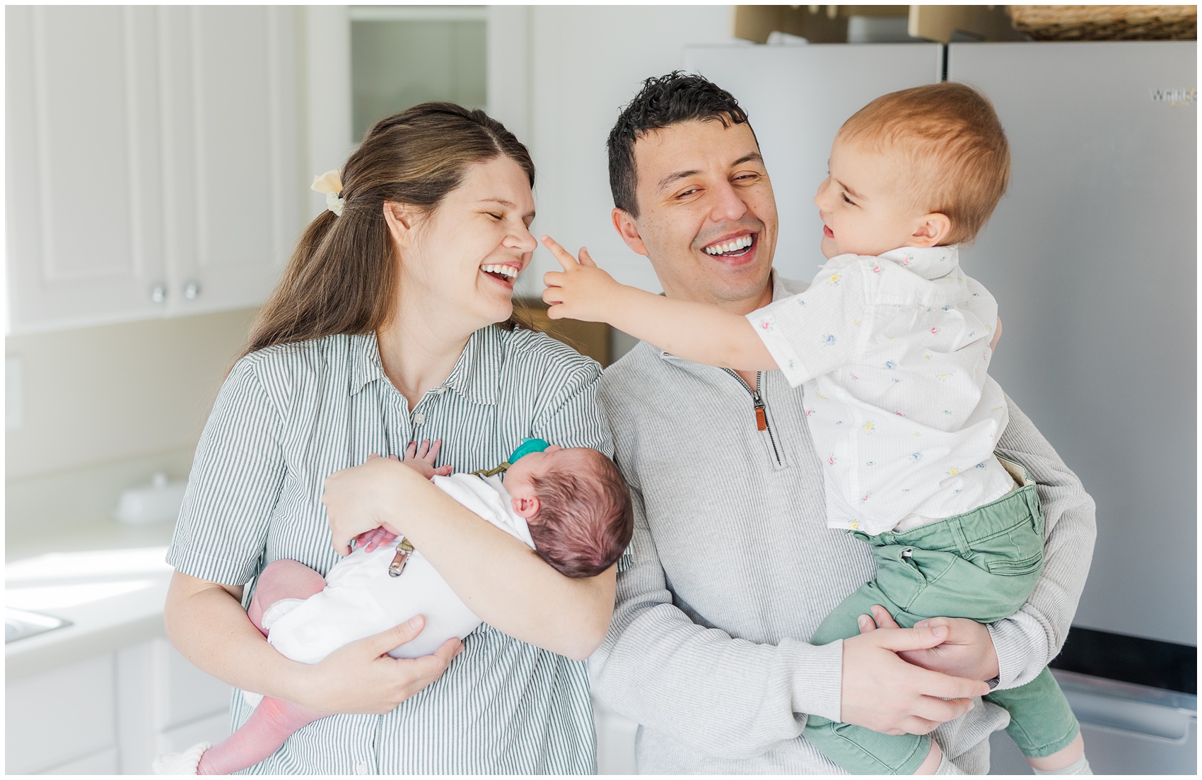 A laughing mom and dad play with their toddler and newborn in a kitchen