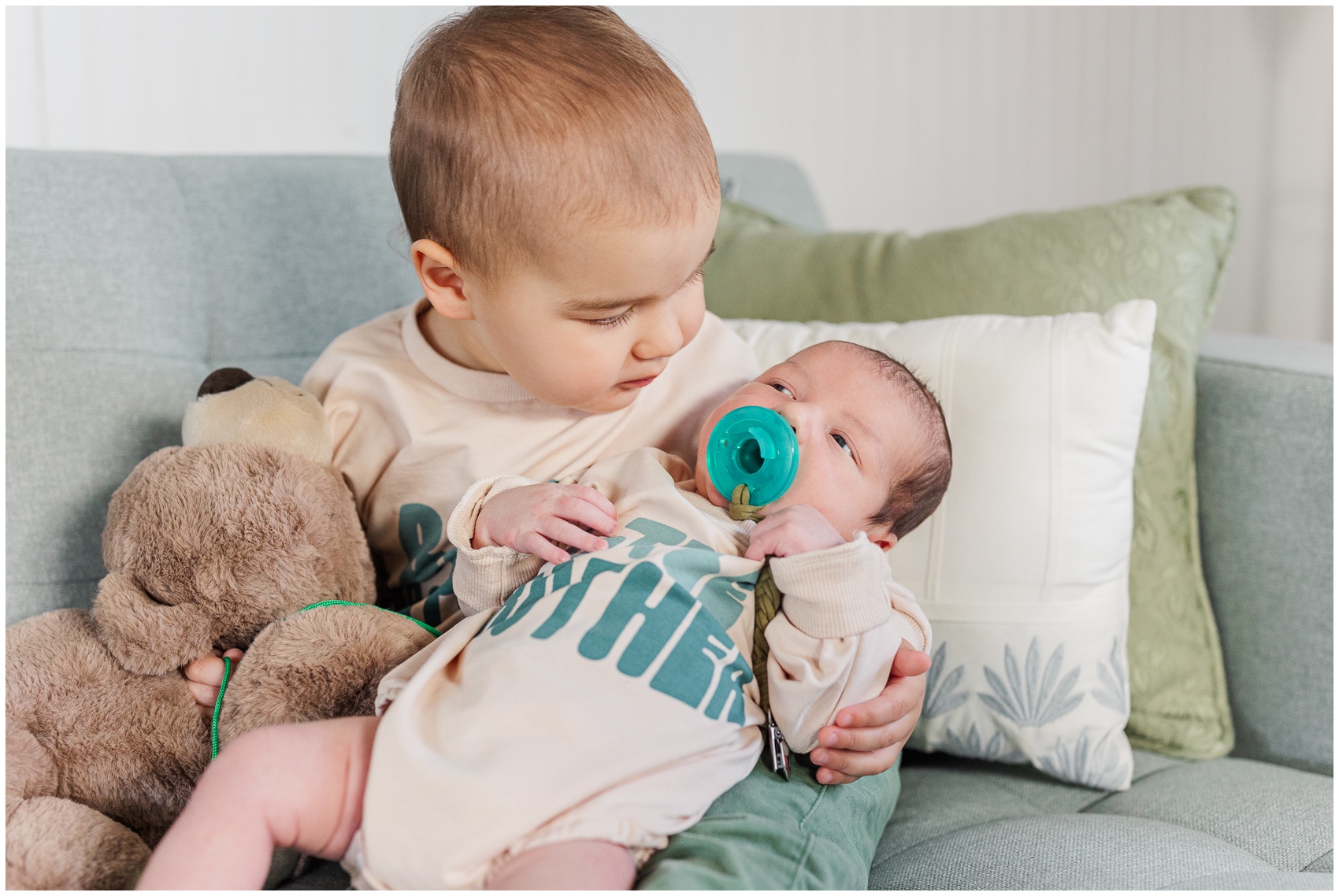 A baby sits on a couch holding a teddy bear and newborn baby brother in his lap after leaving ann arbor birth and family