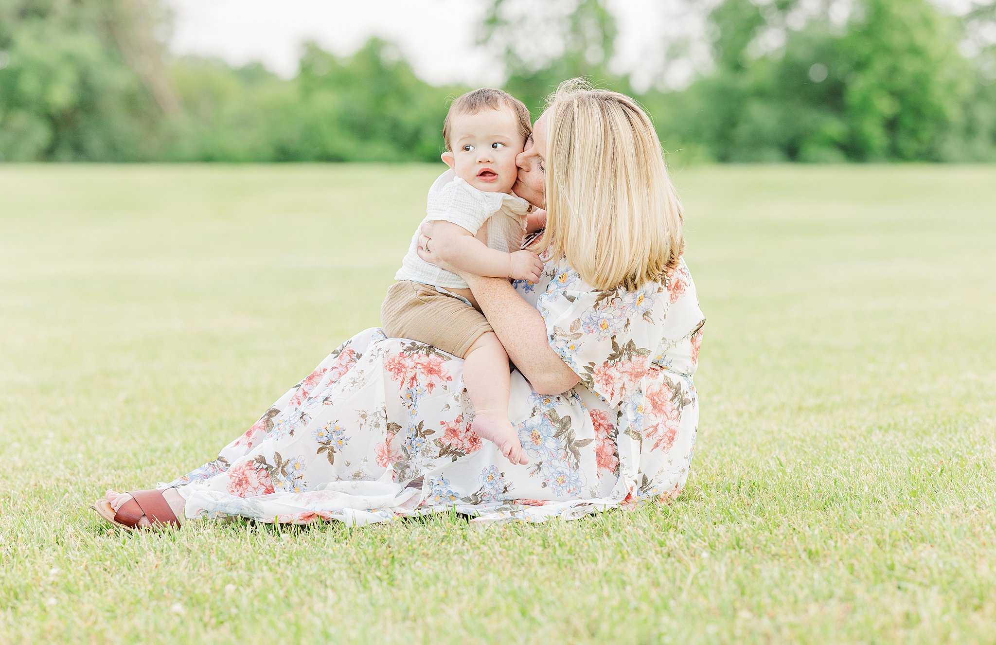 A happy mom kisses her toddler's cheek while he sits in her lap in a park lawn after visiting baby stores in ann arbor