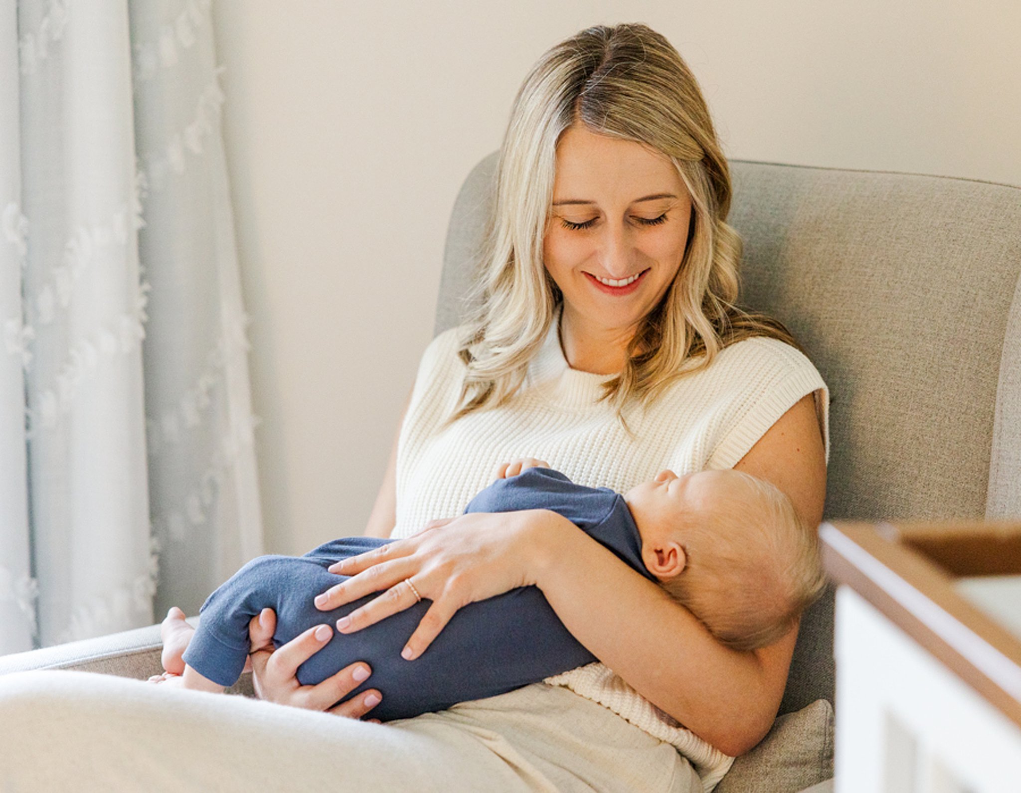 A happy new mom sits in a nursing chair holding her sleeping newborn in a blue onesie after meeting a lactation consultant in ann arbor