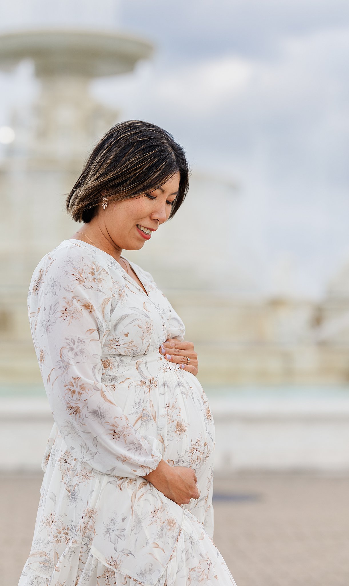 A mother to be in a white floral maternity dress smiles down at her bump in a park after visiting an obgyn in ann arbor