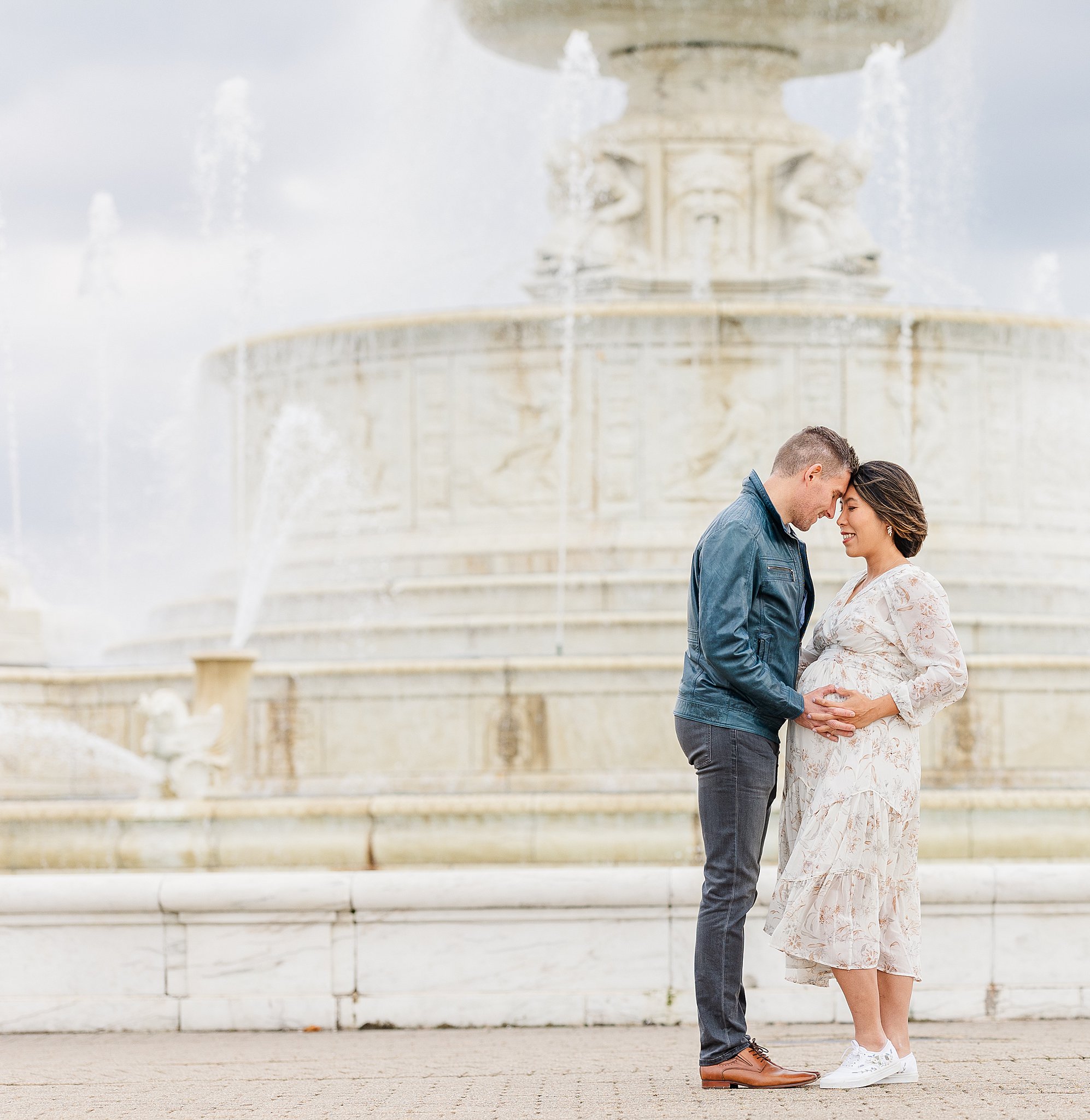 A happy expecting couple hold the bump and touch foreheads by a large fountain after visiting an obgyn in ann arbor