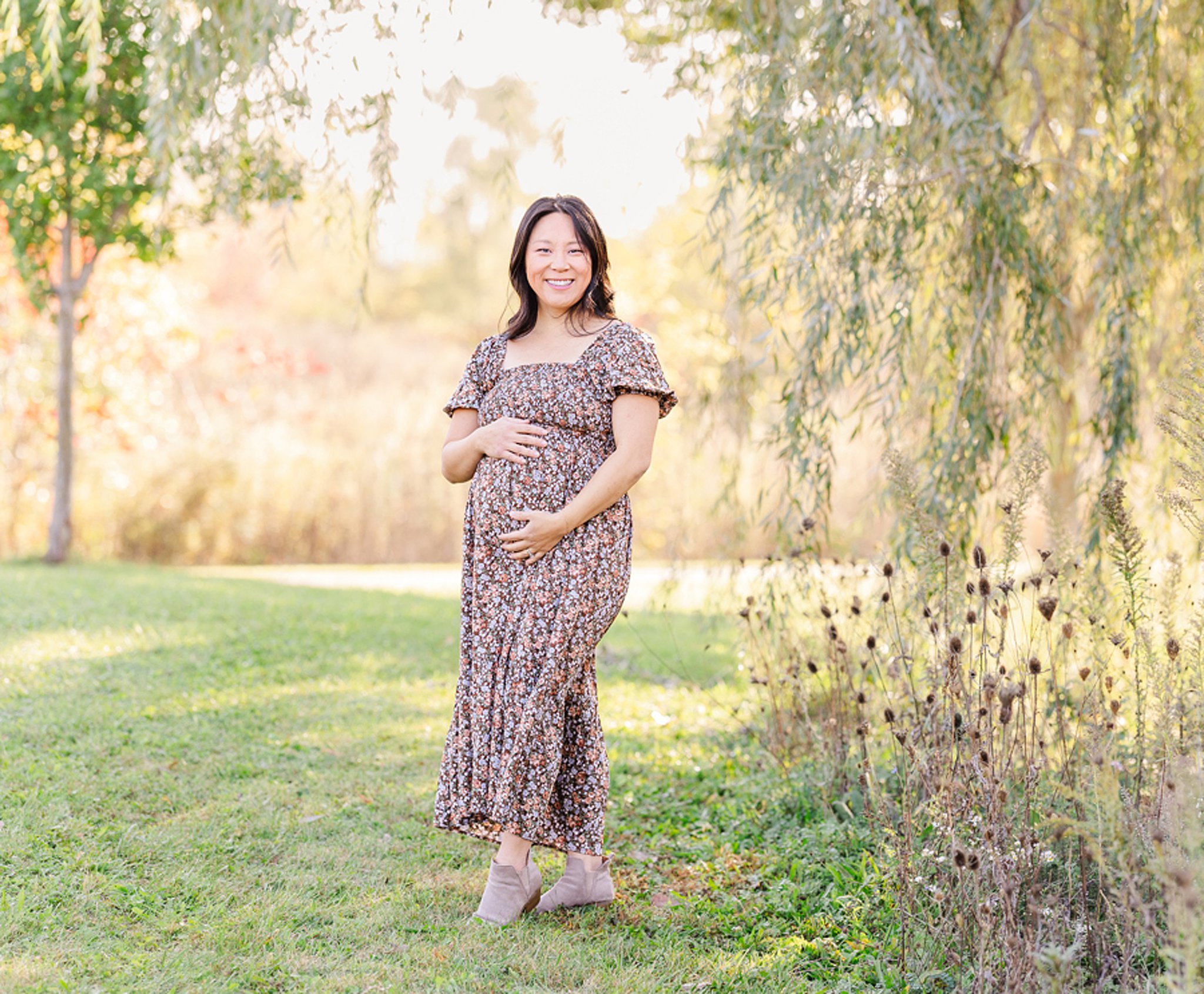 A happy mom to be holds her bump while smiling under a willow tree