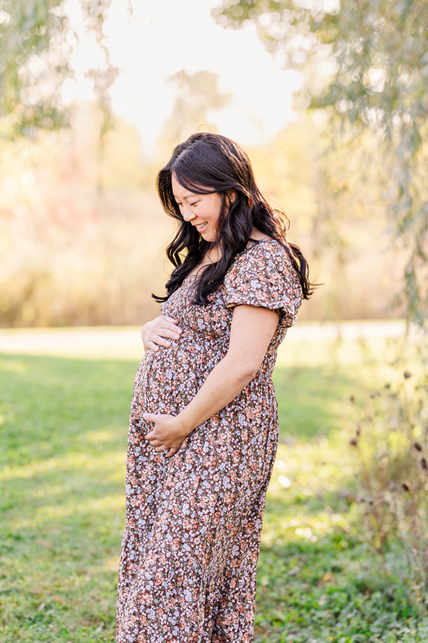 A smiling mom to be looks down at her bump in a floral print dress in a park after visiting peek a bootique