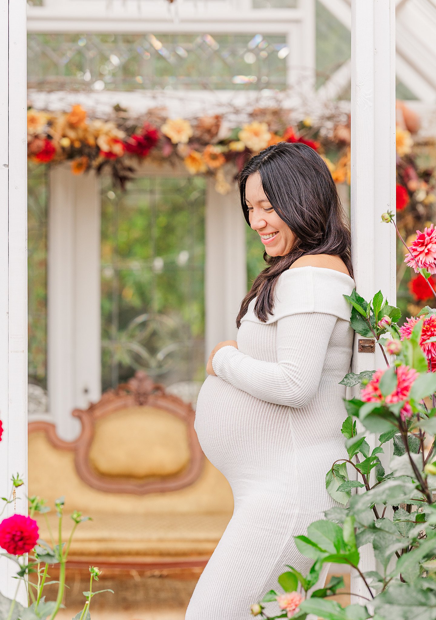 A smiling pregnant woman in a white maternity gown leans on a garden gazebo wall after a prenatal massage in Ann Arbor