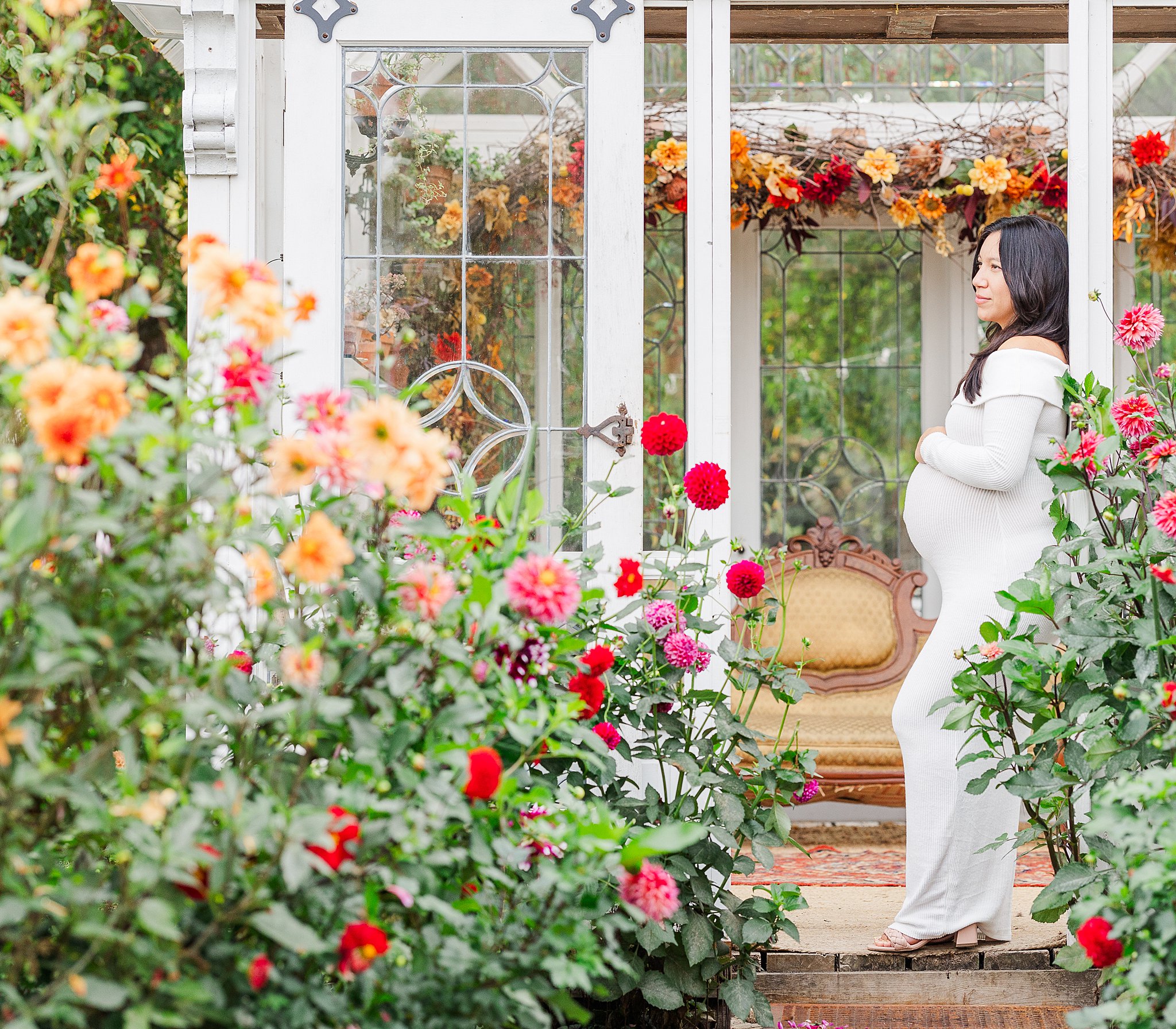 A mom to be in a white maternity gown leans on a greenhouse wall in a garden after a prenatal massage in Ann Arbor