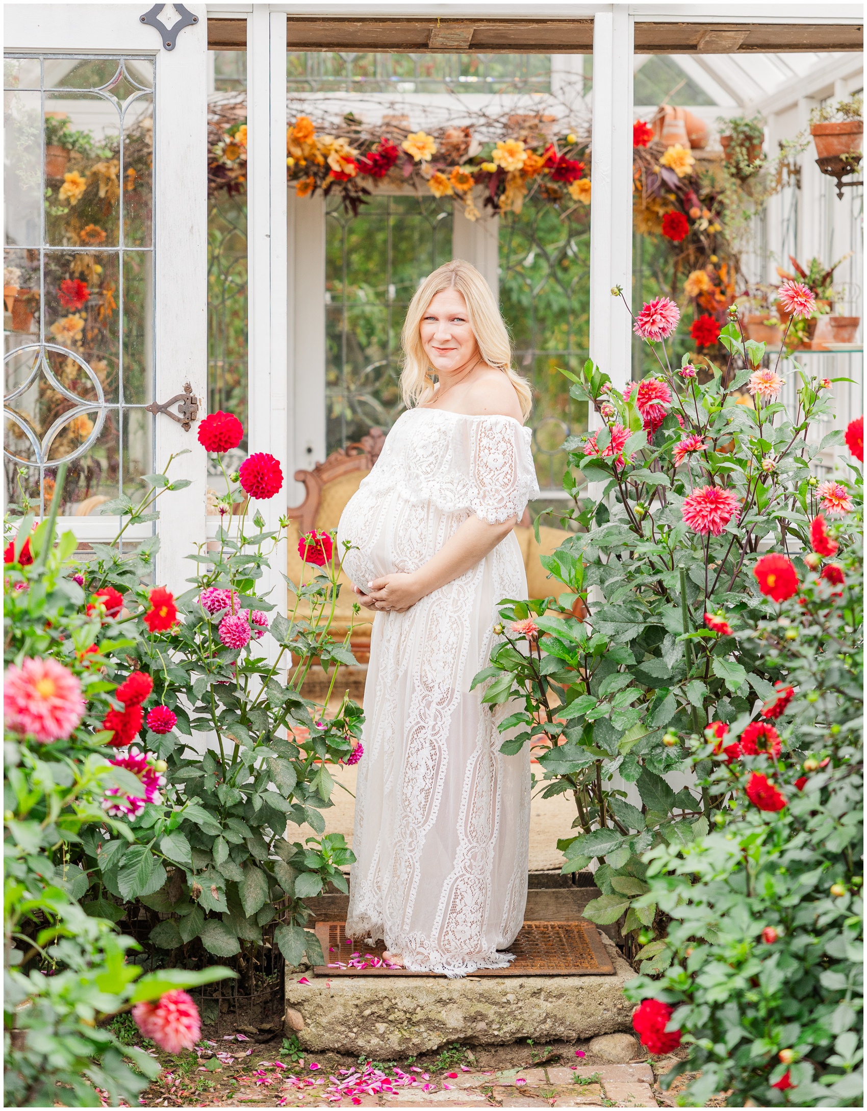 A happy mom to be smiles in a white lace maternity gown while exploring a rose garden