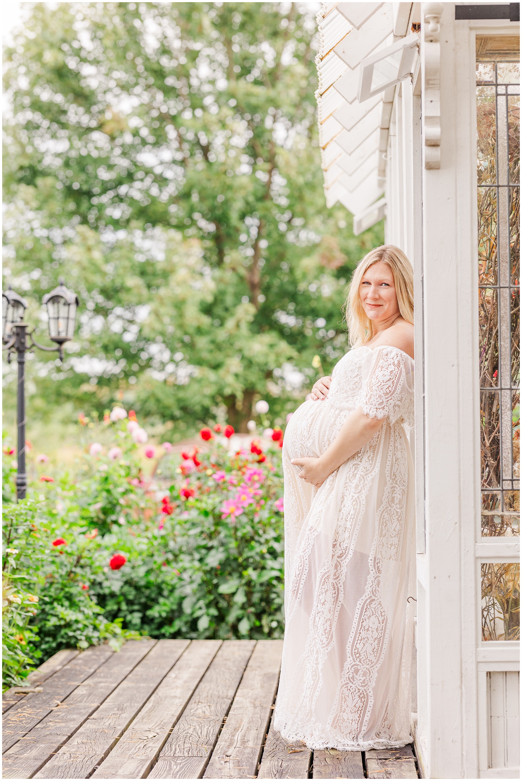 A smiling pregnant woman holds her bump in a lace maternity gown leaning on a garden greenhouse after some prenatal pilates in ann arbor