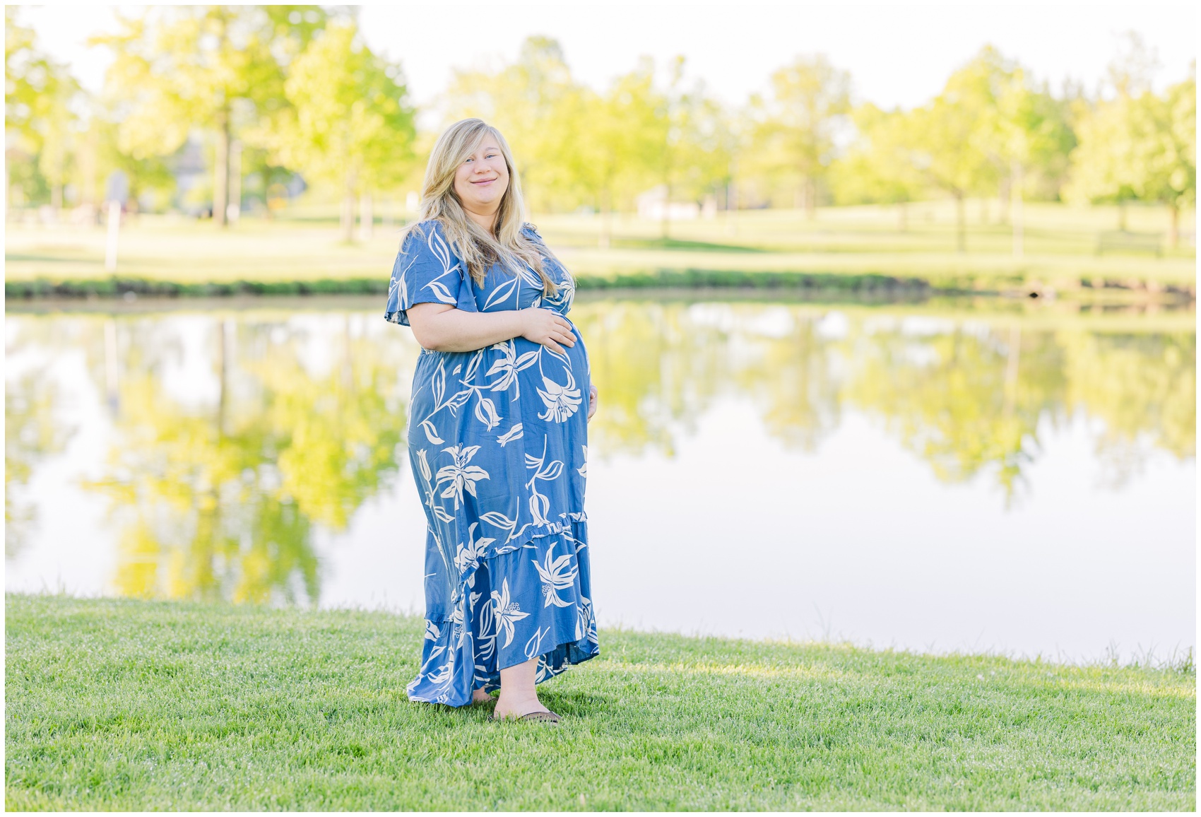 A smiling pregnant woman walks in a park along a pond in a blue floral print dress