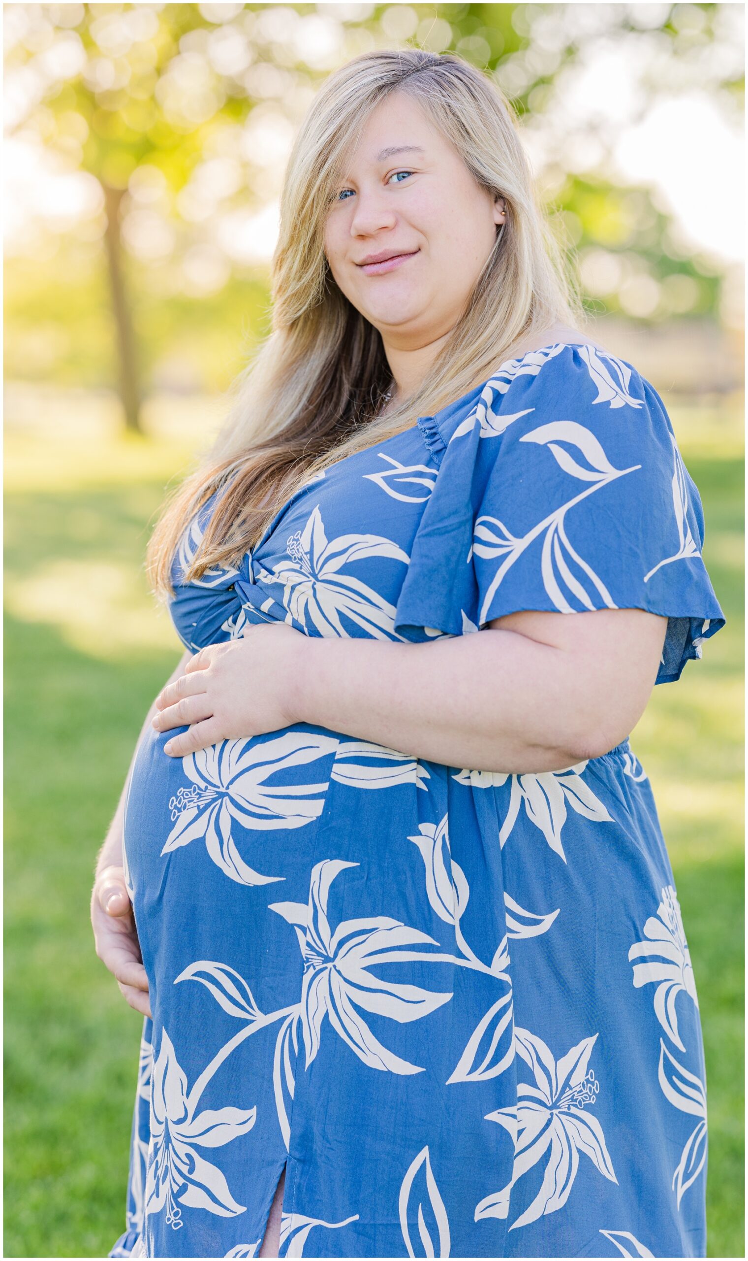 A mom to be in a blue dress smiles while holding her bump in a park after some prenatal yoga in ann arbor