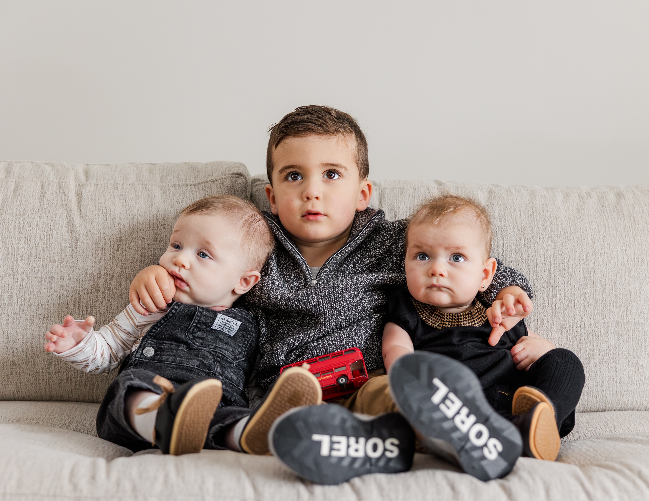 A toddler boy sits on a couch with his arms around two babies