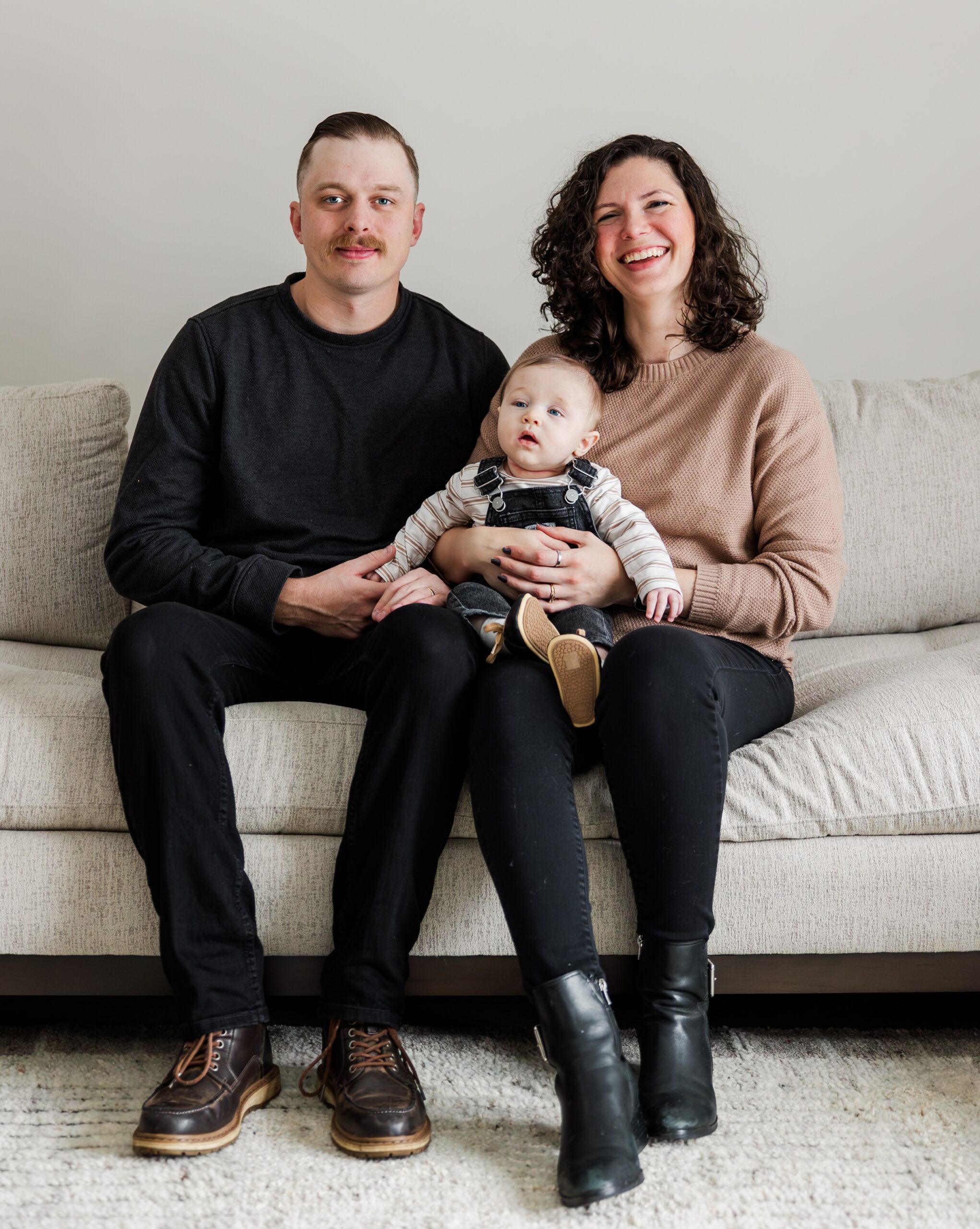 Happy new parents smile on a couch with their baby in mom's lap in overalls after some baby classes in ann arbor