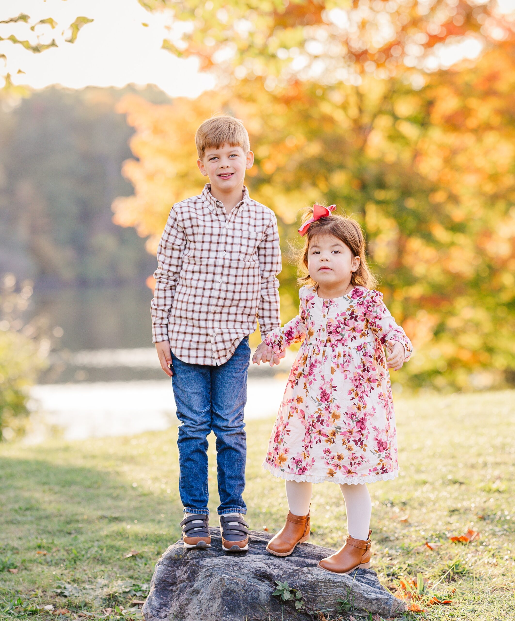 Young brother and sister stand on a rock holding hands in a park at sunset