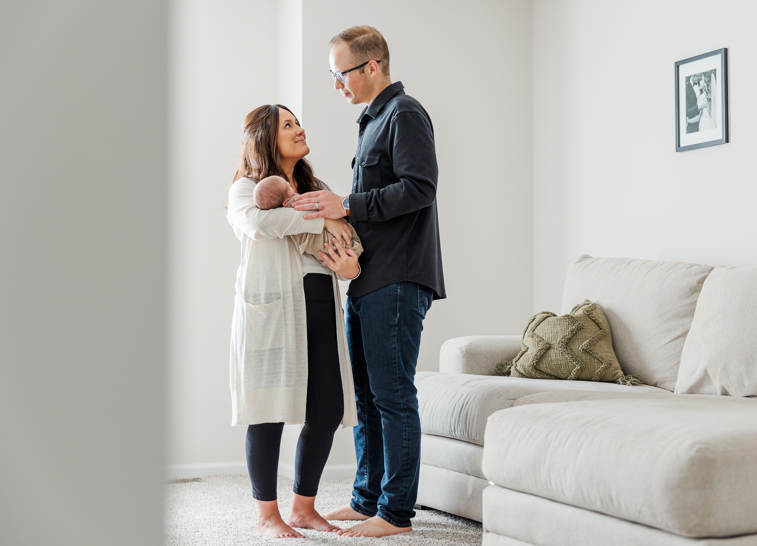 Happy new parents smile at each other while standing in a living room holding their newborn