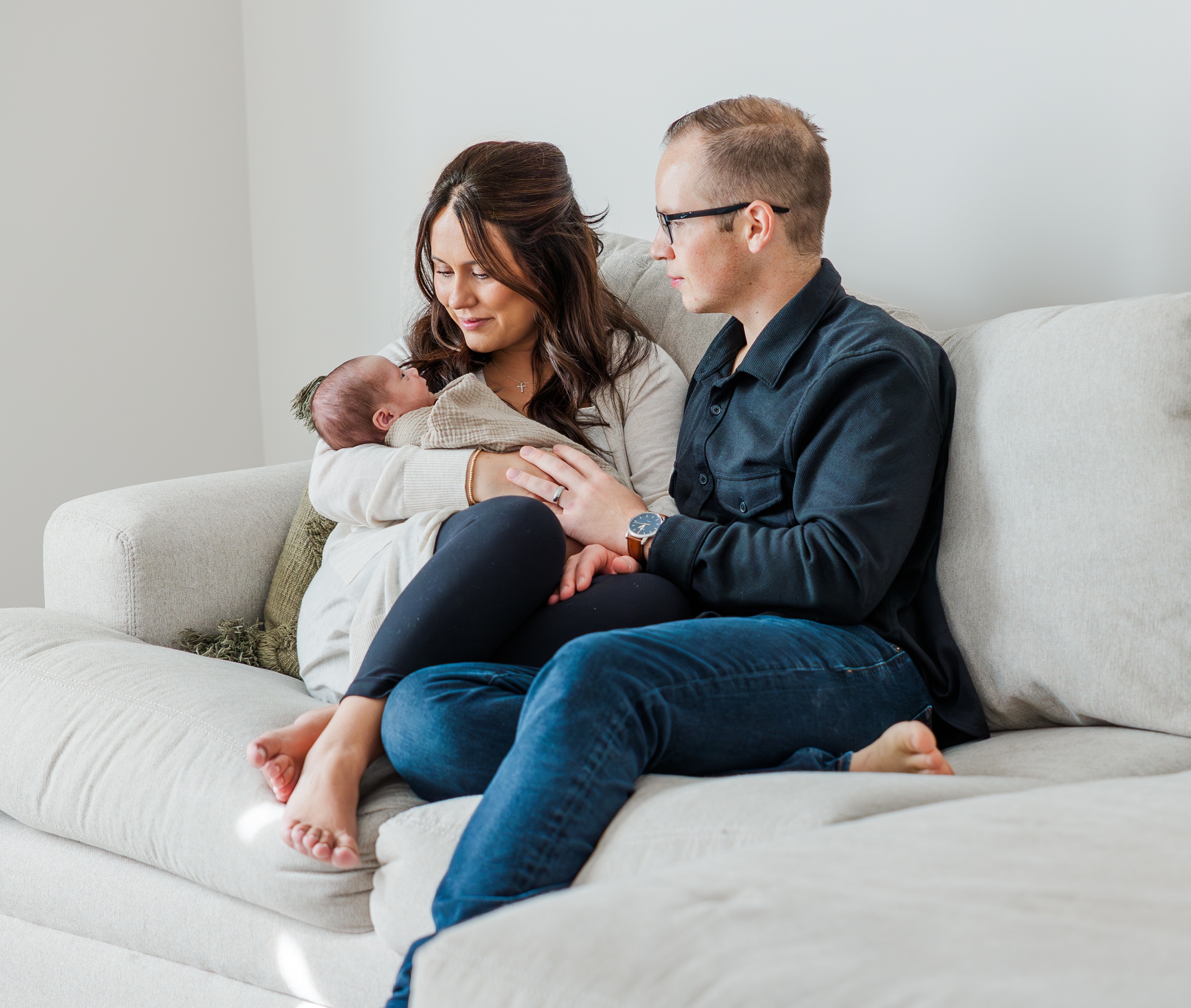 New parents sit on a couch cuddling their sleeping newborn against mom's chest before some pelvic floor therapy in ann arbor