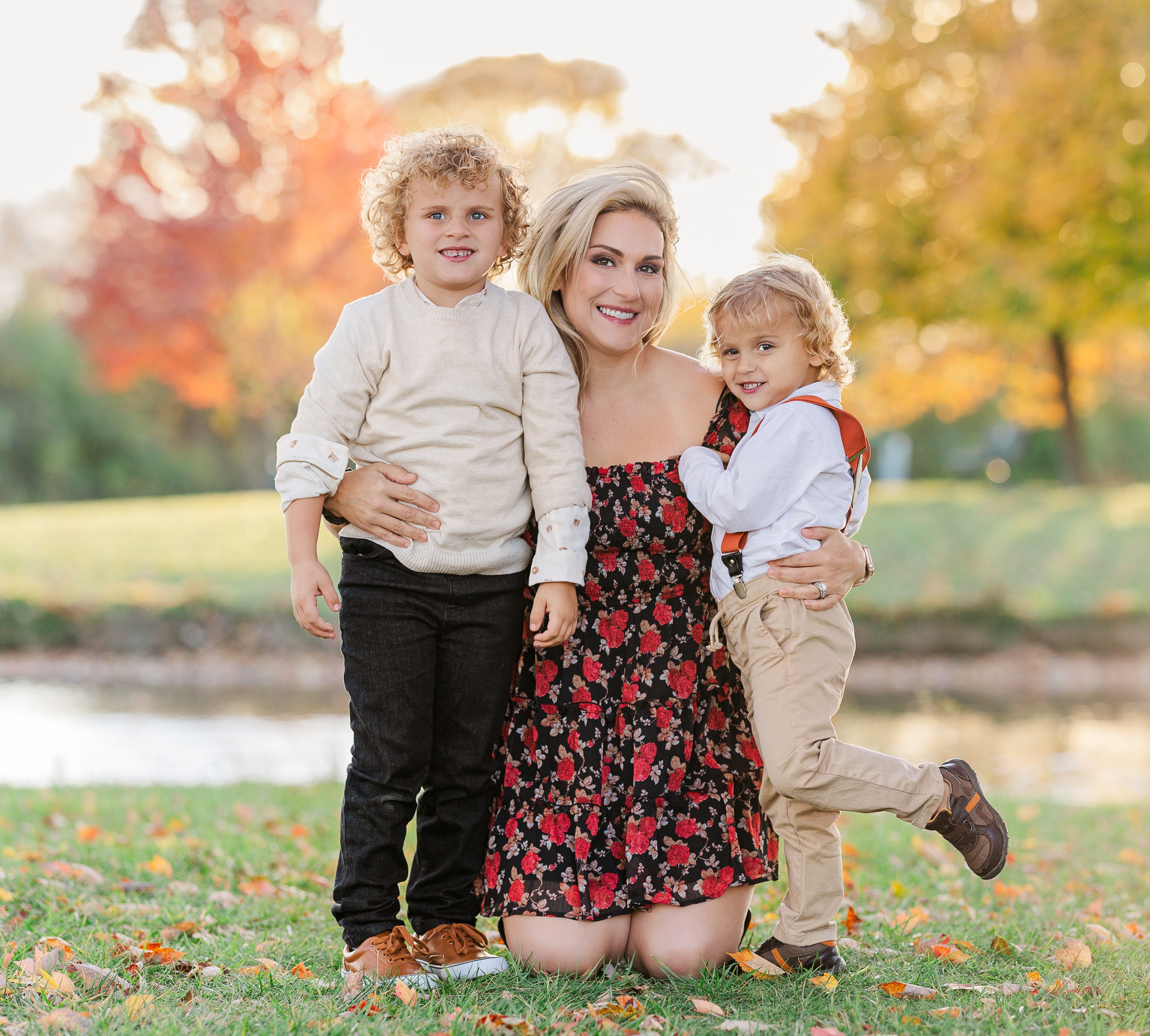 A mom in a red rose print dress kneels in a park hugging her two toddler sons at sunset after some toddler classes in ann arbor